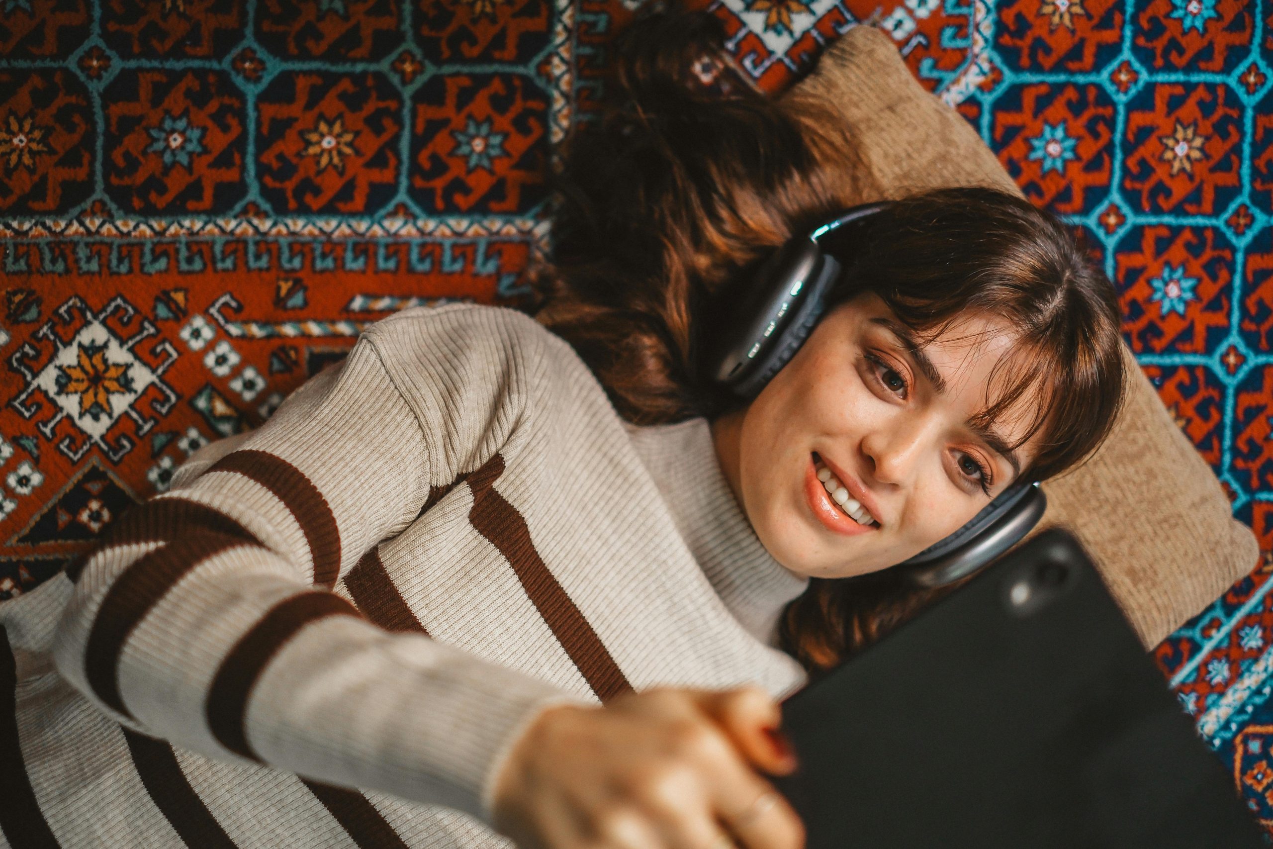 Brunette woman in turtleneck lying on patterned rug with headphones and tablet.